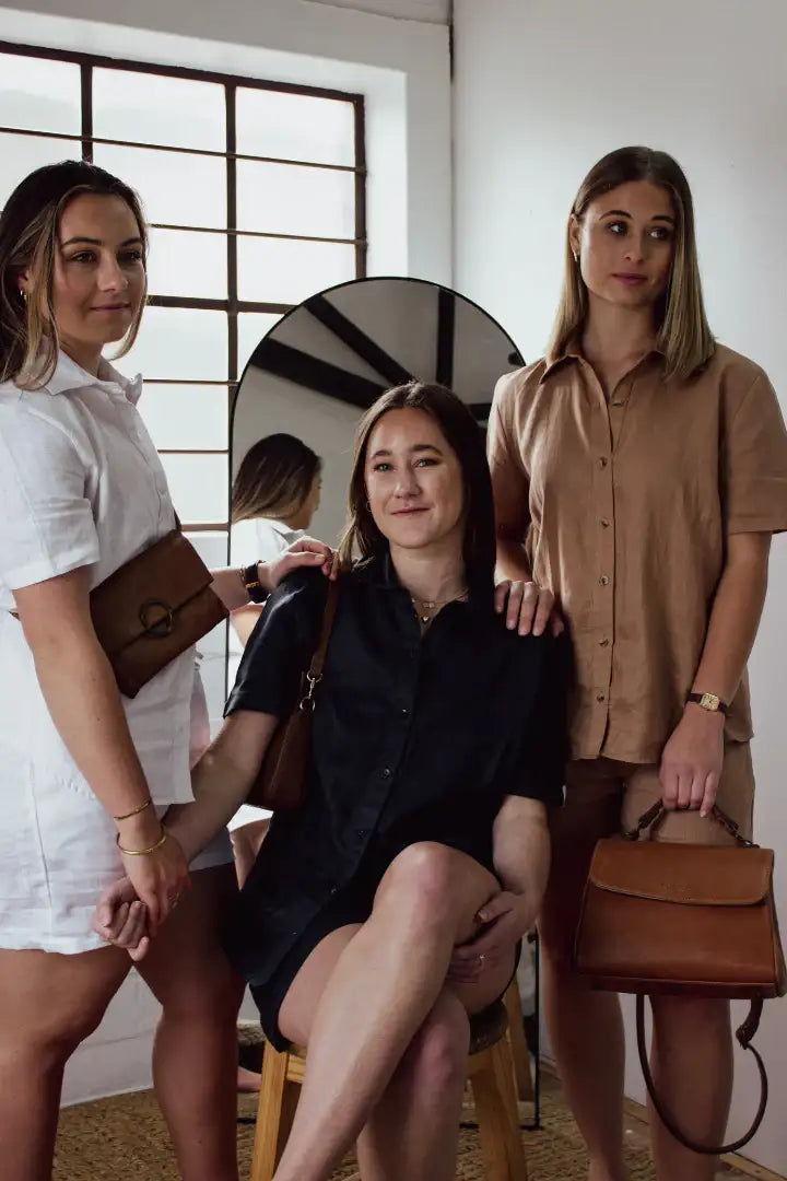 Three women standing indoors, with one sitting on a stool, holding brown handbags in linen clothing