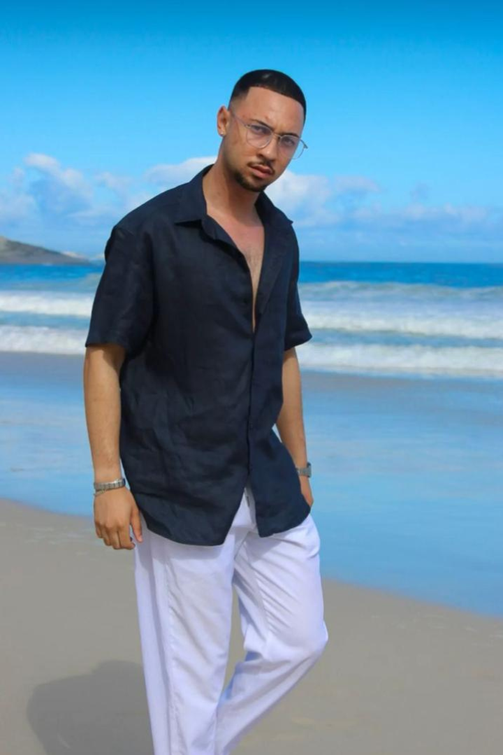 Man standing on a beach wearing a navy shirt and white pants with a blue sky and ocean in the background.