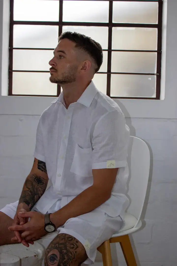 Man sitting on a chair wearing a white linen shirt and shorts in a room with large windows.