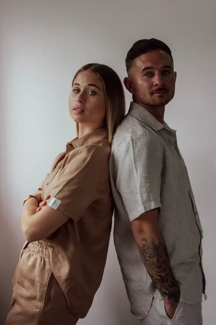 Two people standing back-to-back in 100% Linen outfits against a plain wall.