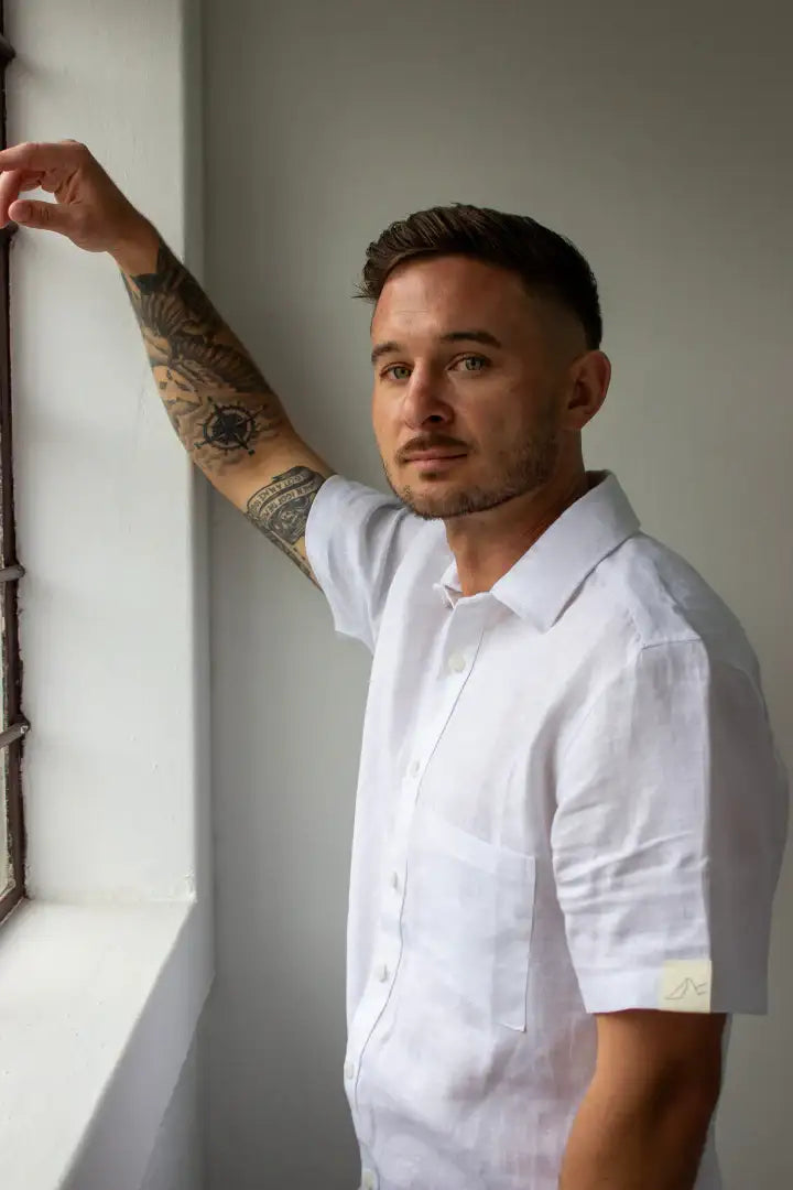 Man wearing a linen white shirt with tattoos on his arm, standing against a neutral wall.
